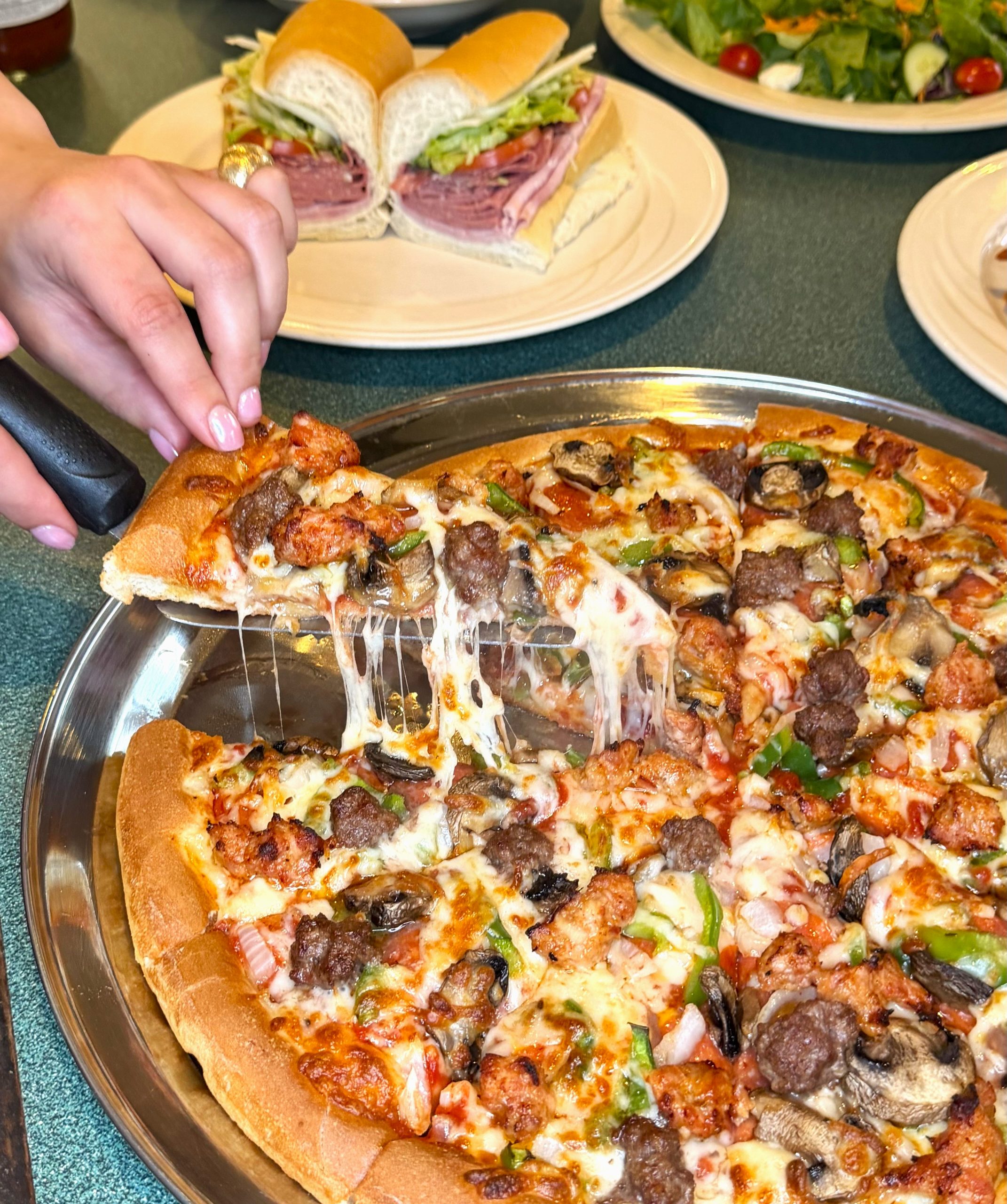 : A hand lifts a slice from a loaded pizza topped with sausage, mushrooms, green peppers, onions, and melted cheese, with long cheese pulls stretching from the pie.